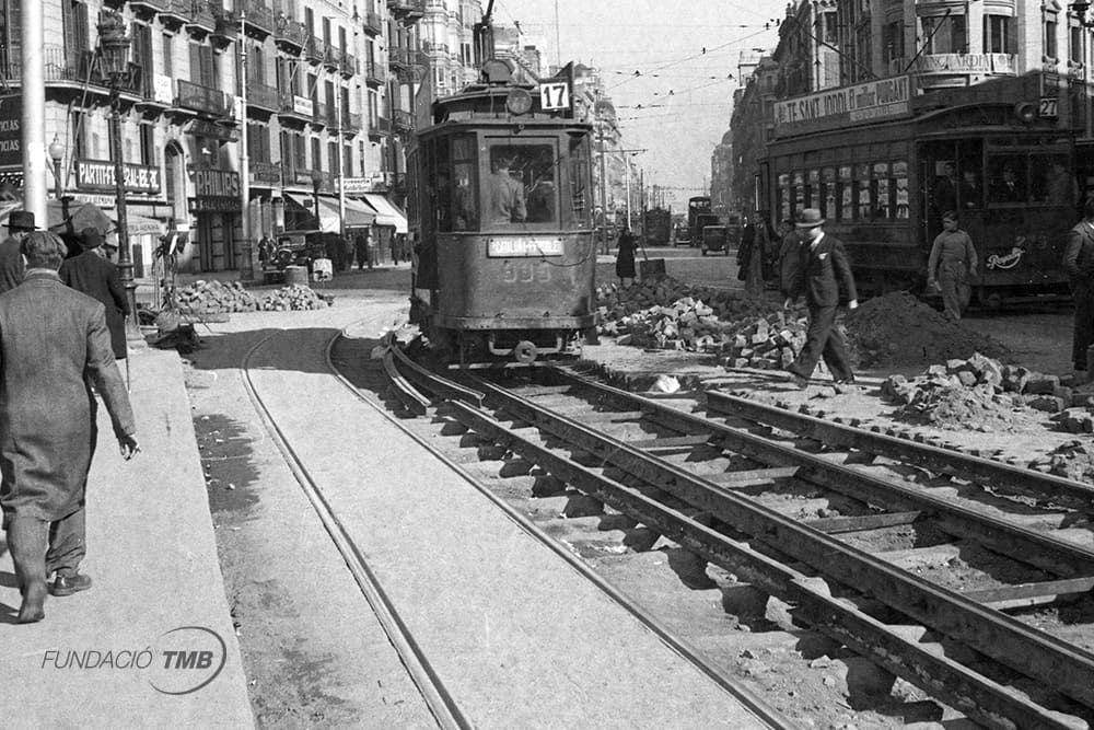 Plaça de la Universitat cantonada Pelai. 1936. Línia 17 Plaça Catalunya- Frederic Soler. Tramvia de via estreta circulant en mig de les obres d’instal·lació de vies amb ample internacional. Per a la renovació de la flota dels antics tramvies va ser necessari substituir la mida de l’ample entre les vies, passant d’un metre a la mida internacional d’1,43 m.